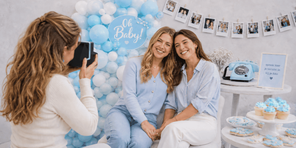 Babyshower fotoshoot met twee lachende vrouwen, polaroidcamera, lichtblauwe EventTelefoon en blauwe feestdecoratie.