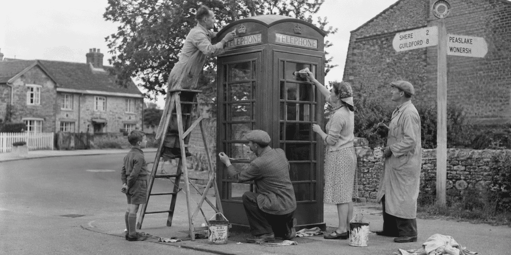 Historisch zwart-witbeeld van dorpsbewoners die gezamenlijk een Britse K6-telefooncel schoonmaken en opnieuw schilderen.