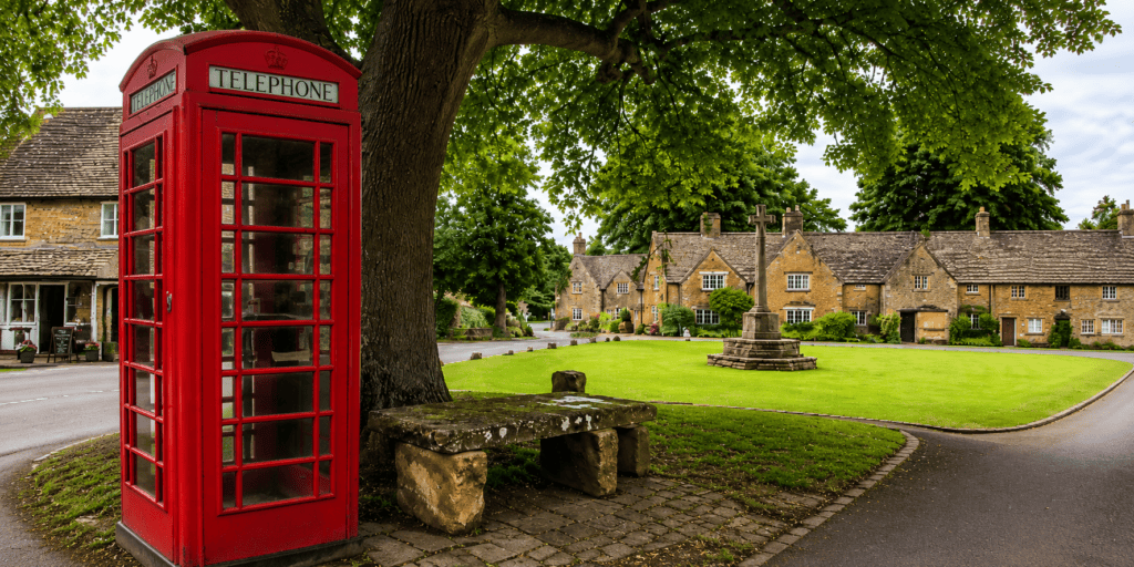 Rode Britse telefooncel onder een grote boom op een sfeervol Engels dorpsplein, met stenen bank, groen gras, monument en historische cottages op de achtergrond.
