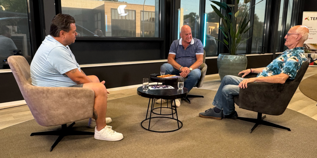 Klaas van den Broek in gesprek met twee materiaalmannen van Tilburg Trappers tijdens zijn interviewprogramma in een moderne zitruimte.