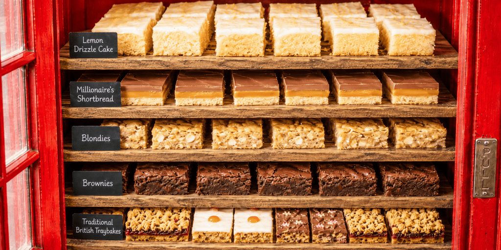 Close-up van lemon drizzle cake, millionaire’s shortbread, blondies, brownies en traditionele Britse traybakes op houten planken in een rode telefooncel.