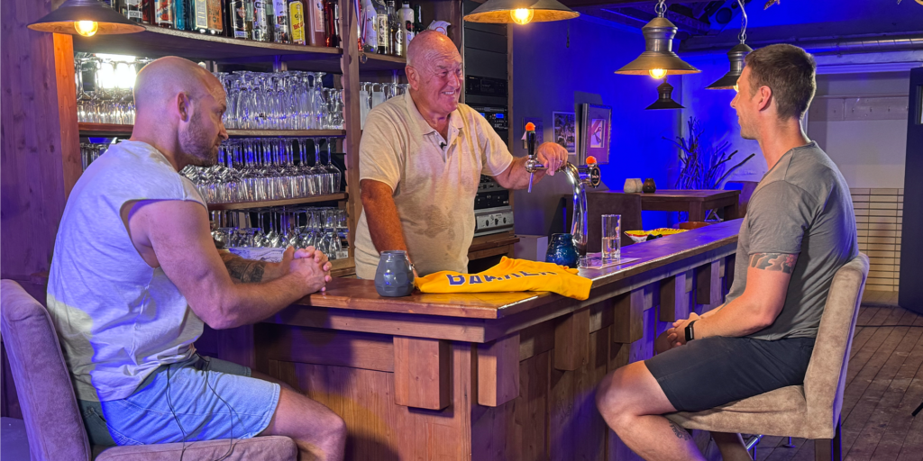 Klas van den Broek, Ivy van den Heuvel en Peter van BIezeb zitten en staan rond een houten bar in een sfeervol verlichte ruimte, in gesprek tussen glazen, flessen en een geel shirt van Guus Bakker op de bar