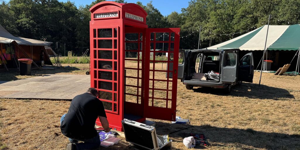 Een rode Britse telefooncel wordt buiten op een evenemententerrein geïnstalleerd. Een persoon werkt gehurkt aan de onderzijde van de telefooncel, met gereedschap en een open koffer op de grond. Op de achtergrond staan tenten en een bestelwagen van eventtelefoon met geopende deuren.