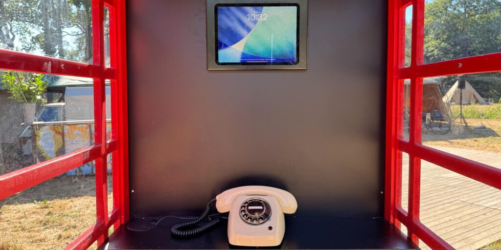 Binnenkant van een rode Britse telefooncel. Op een zwarte plank staat een crèmekleurige eventtelfoon met hoorn en krulsnoer. Boven de telefoon is een beeldscherm in de wand gemonteerd. Door de rode raamkozijnen is buiten een zonnige, groene omgeving zichtbaar.