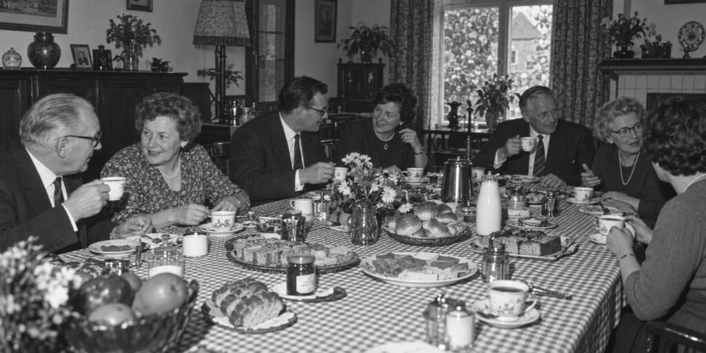 Zwart-witfoto van een Brabantse koffietafel uit de jaren zestig. Acht volwassenen zitten rond een grote tafel met geruit tafelkleed, koffie, broodjes, gebak en beleg. De kamer heeft een huiselijke inrichting met bloemen, servies, gordijnen en houten meubels.