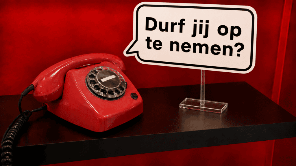 Rode retro EventTelefoon met draaischijf en een spraakballon met de tekst ‘Durf jij op te nemen?’, geplaatst op een transparante plexiglas standaard.