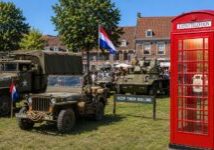 Bevrijdingsevenement met historische militaire voertuigen, Nederlandse vlaggen en bezoekers op een plein. Rechts staat een opvallende rode telefooncel met het opschrift Eventtelefoon’