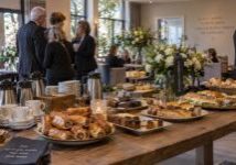 Sfeervolle koffietafel tijdens een afscheid, met een grote houten tafel vol koffie, thee, broodjes, gebak en hartige hapjes. Witte bloemstukken, kaarslicht en teksten over herinnering en afscheid zorgen voor een warme, ingetogen sfeer, terwijl gasten in donkere kleding met elkaar in gesprek zijn.